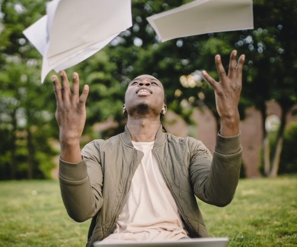 Content black remote worker throwing work papers in air happy to get rid of boring paperwork while sitting in green park with laptop get rid